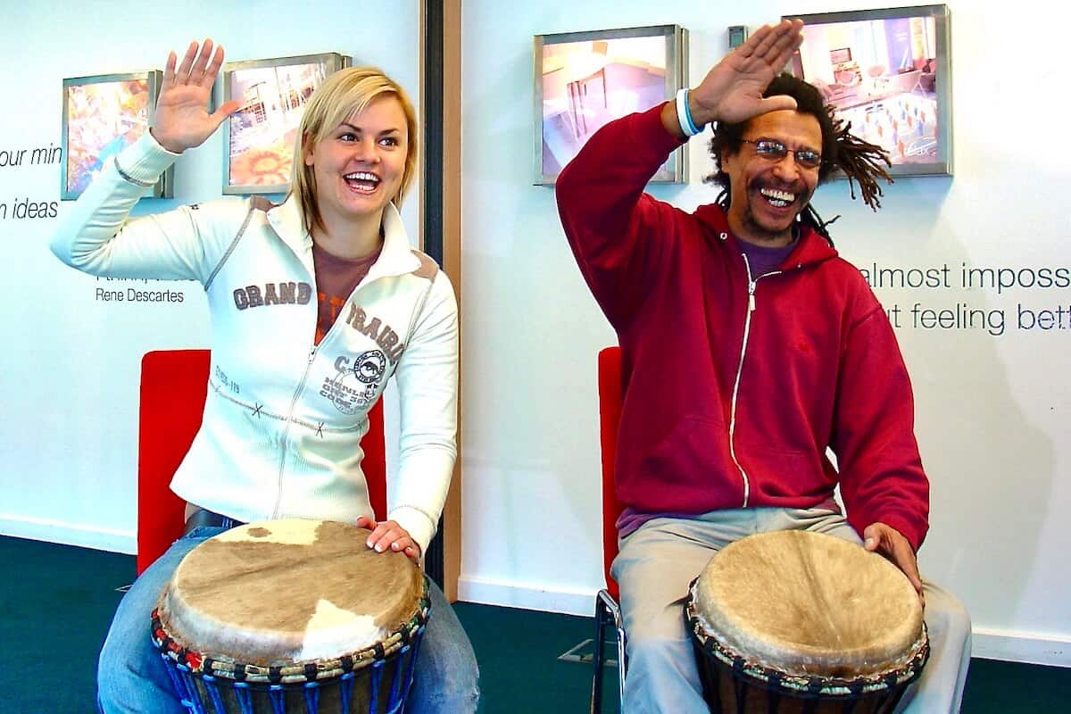 Two smiling participants raising their hands during a corporate drumming workshop, seated with djembes and laughing together – Primal Sound team wellbeing event.