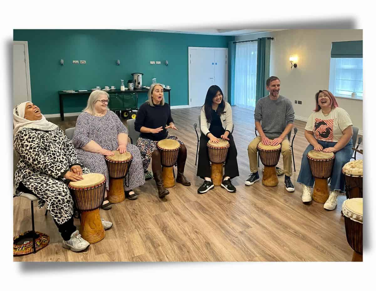 A diverse group of adults laughing and drumming together in a relaxed community wellbeing session.