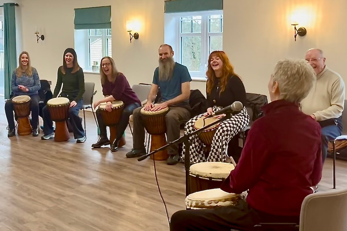 Sarah leading a joyful community drumming session, with participants smiling and playing djembes together in a relaxed circle.